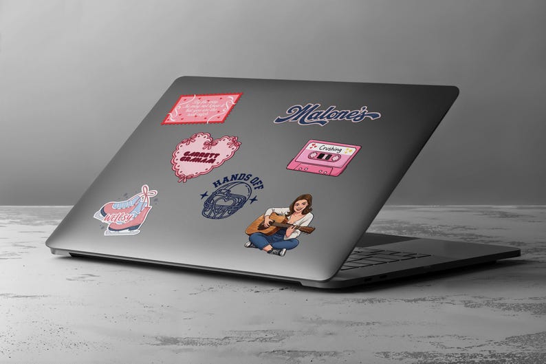 May include: A laptop covered in stickers. Stickers include a pink cassette tape with the word "Crushing", a pink heart with the text "Garrett Graham", and a sticker of a woman playing a guitar. The laptop is on a gray surface.