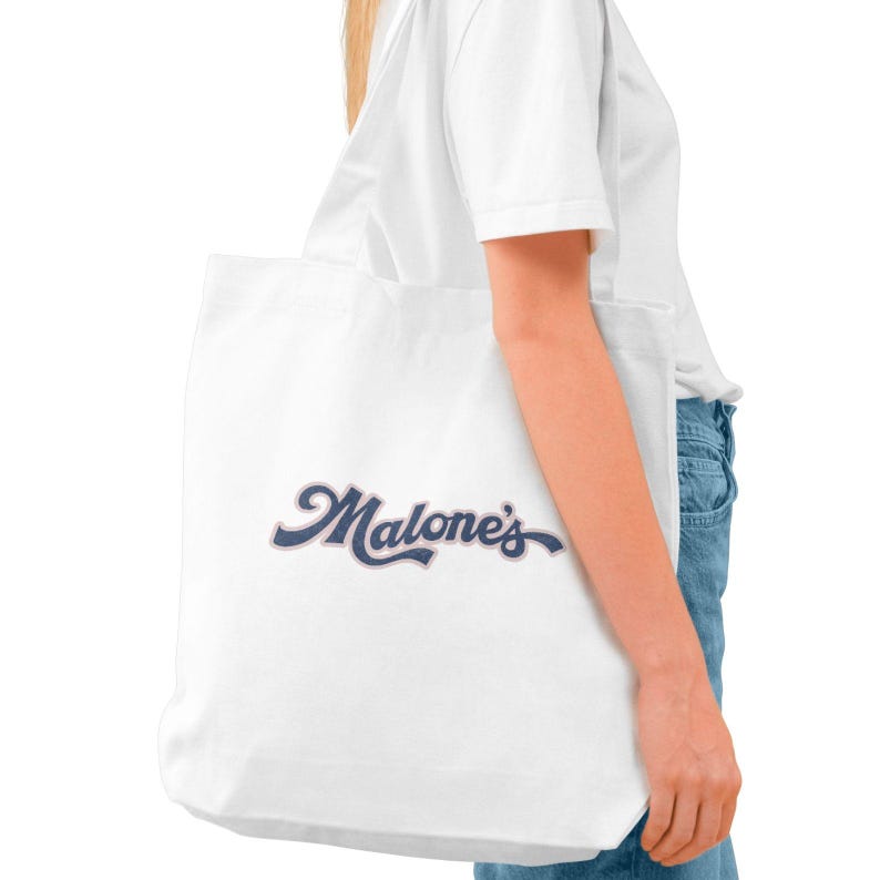 May include: White tote bag with the word "Malone's" in a navy blue script. The bag is made of a light-colored fabric and is being held by a person wearing a white shirt and blue jeans.