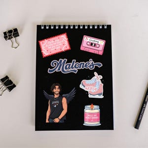 May include: A black spiral-bound notebook with various stickers. Stickers include a pink cassette tape with "Crushing", a pink and blue ice skate, a candle labeled "Smells Like Hockey Romance", and a sticker with the text "By the way, he may not know it but you are the soul catch".