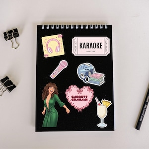 May include: A black spiral-bound notebook with various stickers. Stickers include a pink karaoke ticket, a pink microphone, a woman in a green dress, a pink heart with text, a cocktail, a stack of books, and a pair of headphones.