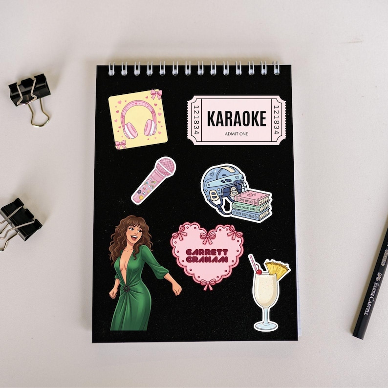 May include: A black spiral-bound notebook with various stickers. Stickers include a pink karaoke ticket, a pink microphone, a woman in a green dress, a pink heart with text, a cocktail, a stack of books, and a pair of headphones.