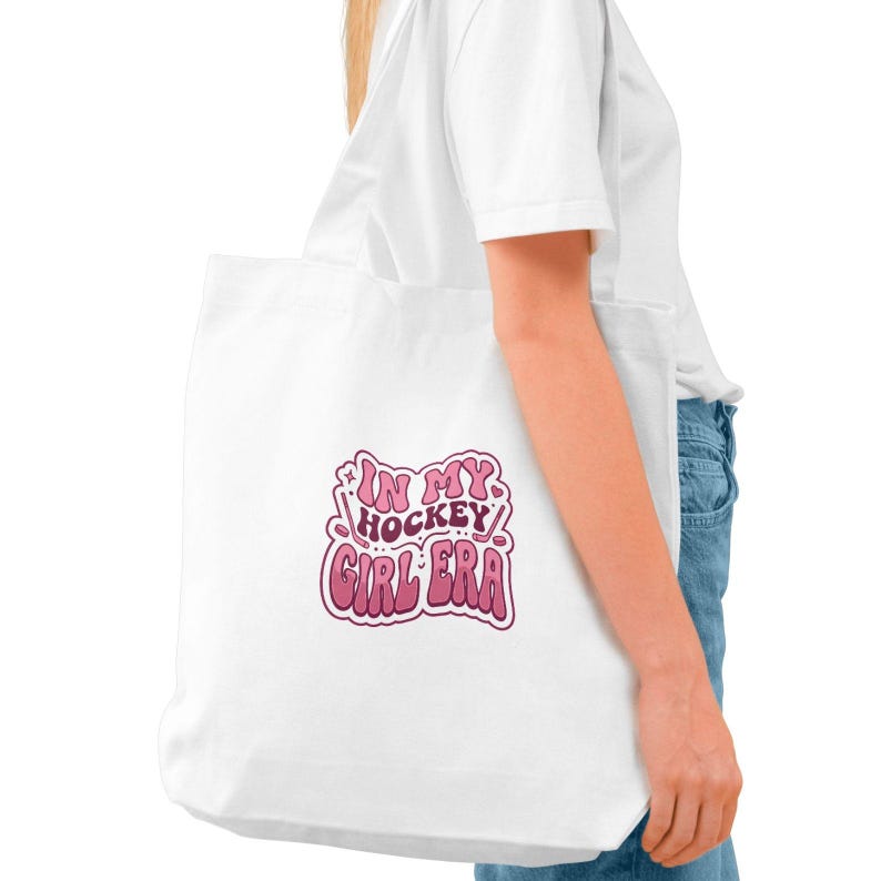 May include: White tote bag with the text "IN MY HOCKEY GIRL ERA" in pink and purple. The bag is made of a soft material and is being held by a person.