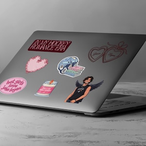 May include: A laptop covered in stickers. Stickers include a pink heart, a hockey helmet, a candle, a winged figure, and text that reads "IN MY HOCKEY ROMANCE ERA" and "hot girls love John Logan."