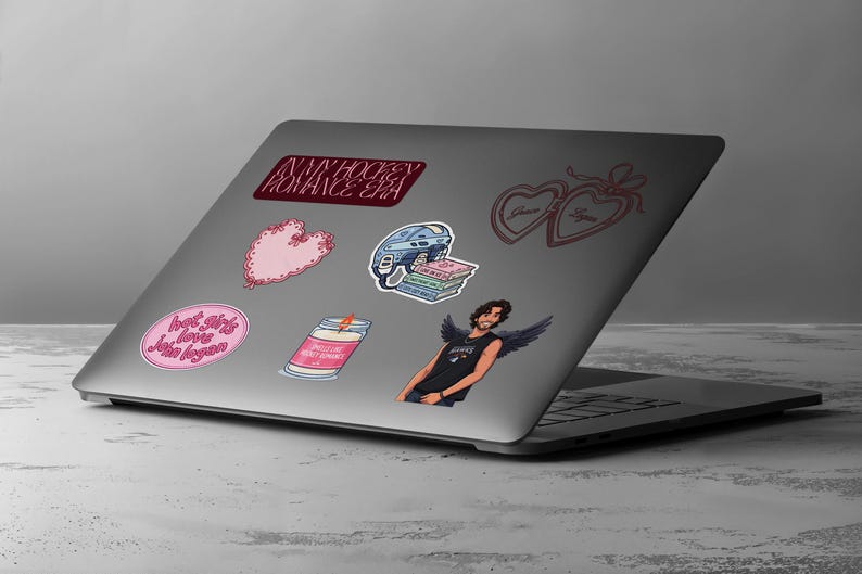 May include: A laptop covered in stickers. Stickers include a pink heart, a hockey helmet, a candle, a winged figure, and text that reads "IN MY HOCKEY ROMANCE ERA" and "hot girls love John Logan."
