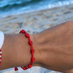 Puede incluir: Una pulsera roja anudada se lleva en la muñeca. La pulsera está hecha de cordón rojo y presenta varios nudos. El fondo muestra una playa con arena y olas del océano.