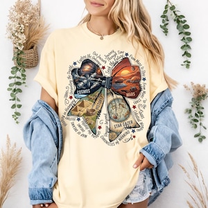 Puede incluir: Una camiseta amarilla clara con un gran diseño de lazo que incorpora imágenes espaciales e históricas. El lazo incluye las palabras "Cosmos", "Mars" y "Star Gazer". Alrededor del lazo hay frases como "Explore the Unknown" y "Chronicles of Mankind".