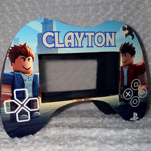 Könnte beinhalten: Ein individuell gestalteter, Controller-förmiger Rahmen mit dem Namen "CLAYTON" in blauen Buchstaben. Der Rahmen zeigt Cartoon-Charaktere und Gamecontroller-Symbole. Der Hintergrund zeigt eine Stadtlandschaft und Himmel.