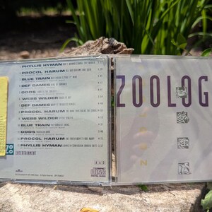 May include: A clear plastic CD case, open, displaying the disc and album art. The album cover reads "ZOOLOGY" in purple text. The tracklist is visible on the left side. A yellow sticker is affixed to the case.