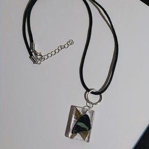 May include: A black cord necklace with a rectangular pendant. The pendant is clear and contains a preserved butterfly wing with black and teal coloring. The necklace has a silver-colored clasp and chain.