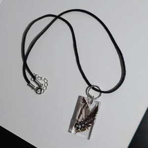 May include: A rectangular pendant necklace with a black cord. The pendant is clear, encasing dried leaves and small, dark blue beads. The necklace has a silver-colored clasp.