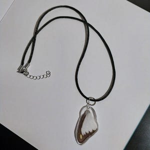 May include: A black cord necklace with a pendant featuring a butterfly wing encased in clear resin. The wing displays white, brown, and yellow hues. The necklace has a silver-colored clasp and a small chain for adjustable length.
