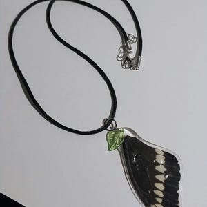 May include: A black cord necklace with a pendant featuring a preserved butterfly wing. The wing is black with white markings and is encased in clear resin. A small green leaf charm is attached to the pendant.