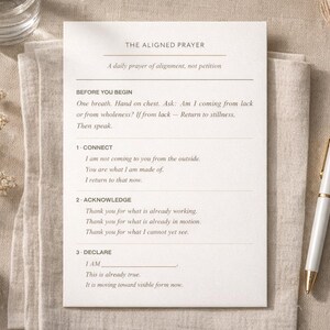 Op de afbeelding: Een bedrukte kaart getiteld "THE ALIGNED PRAYER" met tekst over dagelijkse afstemming. Een witte pen met gouden accenten ligt naast de kaart. De kaart ligt op een beige linnen doek, met gedroogde bloemen en een steen in de buurt.