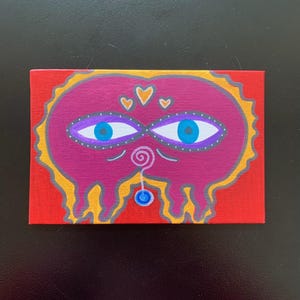 May include: A hand-painted canvas with a pink and purple abstract face with blue eyes, a yellow swirl, and a blue bead. The background is red.