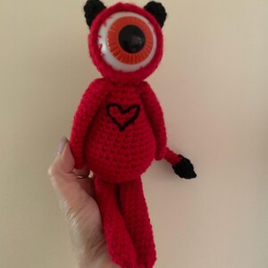 May include: A handmade red crochet doll with a large eye, black ears, and a black heart on its chest. The doll has a black tail and is held up by a hand. The eye is white, orange, and black.