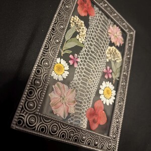 Puede incluir: Un marco rectangular plateado con un dise&ntilde;o de piel de serpiente y flores secas. Las flores son de color rosa, rojo, amarillo y blanco, sobre un fondo oscuro. El marco tiene un patr&oacute;n decorativo en espiral.
