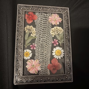 May include: A rectangular silver picture frame with a decorative border. Inside, a dark background features a snakeskin pattern and an arrangement of dried flowers in red, pink, white, and yellow.