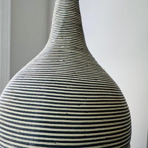 May include: A tall, bulbous vase featuring a narrow neck and a wide, rounded base. The entire exterior is covered in a tight, horizontal pattern of thin, alternating black and off-white stripes, creating a textured, coiled appearance throughout.