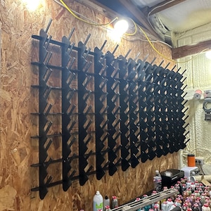 Puede incluir: Un gran estante negro de metal para carretes de hilo montado en una pared de paneles de madera. El estante está lleno de numerosos carretes de hilo de varios colores. El taller incluye luces brillantes y una mesa con más carretes.