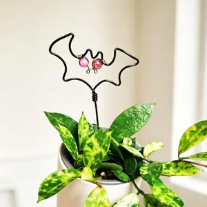 May include: A black wire plant stake shaped like a bat silhouette with two small pink beads positioned as eyes. The wire bat is mounted on a single vertical wire stem that extends downward into a potted plant with variegated green leaves.