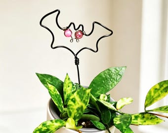 Pode incluir: Um suporte para plantas em arame preto com o formato de uma silhueta de morcego e duas pequenas contas cor-de-rosa como olhos. O morcego de arame está montado numa haste vertical inserida num vaso com folhas verdes variegadas.