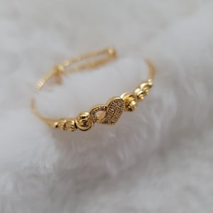 May include: A gold-coloured bangle bracelet featuring a heart-shaped charm. The heart is adorned with small, clear gemstones. The bracelet has a delicate design with small, round beads and a clasp for adjusting the size. It rests on a white, fluffy surface.