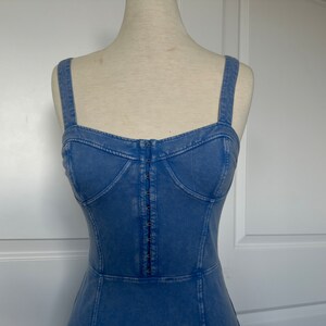 May include: A light blue denim dress with a fitted bodice and a straight skirt. The dress has a button-up front and thin straps.
