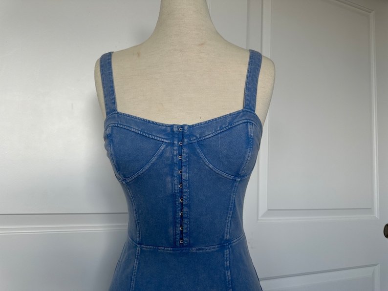 May include: A light blue denim dress with a fitted bodice and a straight skirt. The dress has a button-up front and thin straps.