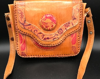 May include: A tan leather shoulder bag featuring a flap closure with a circular red and tan embossed emblem. The bag is decorated with intricate floral and paisley patterns in red and brown, finished with whip-stitched edges and a long adjustable strap.
