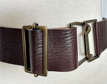 May include: A brown leather belt with a brass buckle. The buckle has a rectangular shape with a smaller rectangular opening.
