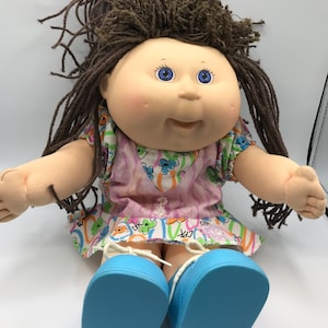 Könnte beinhalten: Eine Puppe mit braunen Wollhaaren, blauen Augen und Grübchen in den Wangen. Sie trägt ein kurzärmeliges Kleid mit einem bunten Muster aus Blumen, Herzen und dem Text CPK. Die Puppe hat hellblaue Schuhe mit weißen Schnürsenkeln an der Vorderseite.