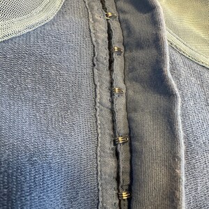 May include: A close-up of a blue fabric garment with a row of metal hooks and eyes along the center seam.
