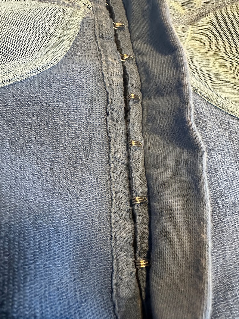 May include: A close-up of a blue fabric garment with a row of metal hooks and eyes along the center seam.