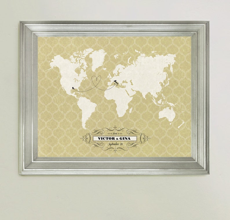 May include: A framed world map print with a heart-shaped line connecting two locations. The map is on a beige background with a decorative pattern. The text "VICTOR & GINA" and "September 20" is printed below the map.