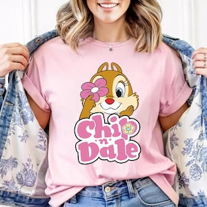 May include: A light pink t-shirt featuring a cartoon image of Chip from Chip 'n' Dale holding a pink flower over one eye. The words "Chip 'n' Dale" are in pink and white bubble letters. The model is wearing a denim jacket.