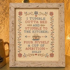 May include: A framed cross-stitch sampler featuring the text I TUMBLE OUTTA BED AND STUMBLE TO THE KITCHEN; POUR MYSELF A CUP OF AMBITION - DOLLY in red and blue thread. The design includes a coffee cup icon and floral borders in a distressed white frame.