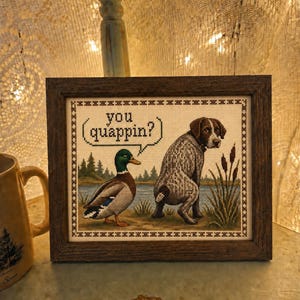 May include: A framed cross-stitch embroidery featuring a mallard duck and a speckled hunting dog in a landscape with a lake and trees. The duck has a speech bubble containing the text 'you quappin?'. The piece is enclosed in a dark wood frame.