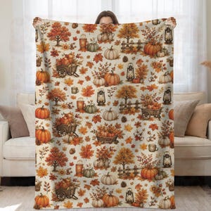 May include: A cream-colored throw blanket featuring a repeating autumn-themed pattern of orange pumpkins, brown wooden wheelbarrows, rustic lanterns, fences, fall foliage, pinecones, and mugs filled with warm beverages on a light background.