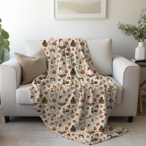 Peut inclure: Un plaid de couleur crème orné d'un motif répétitif de fantômes blancs portant des bonnets et des écharpes en tricot, des citrouilles orange, des piles de livres, des lanternes, des tasses de boissons chaudes et des feuilles d'automne.