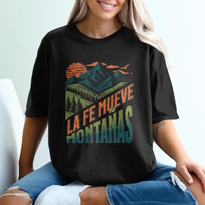 May include: Black t-shirt with a colorful graphic of mountains, a sunset, and the text "LA FE MUEVE MONTAÑAS" in orange and green. The shirt is a crew neck and has short sleeves.