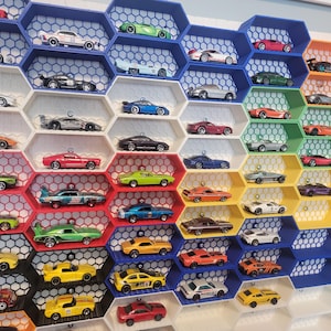 Peut inclure: Collection de voitures miniatures colorées exposées dans une étagère en forme de nid d'abeille. Les voitures sont de différentes couleurs, notamment rouge, bleu, vert, jaune et blanc. L'étagère est en plastique et est fixée au mur.