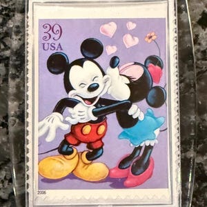 May include: A clear keychain featuring a postage stamp with Mickey Mouse and Minnie Mouse embracing. The stamp has a purple background with hearts and a flower. The stamp reads "39 USA" and "2006". Mickey wears red shorts and yellow shoes.