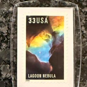May include: Clear acrylic keychain featuring a 33 USA postage stamp. The stamp depicts the Lagoon Nebula with vibrant colors of yellow, blue, and brown. The text "LAGOON NEBULA" and "2000" are visible.