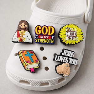 Könnte beinhalten: Weißer Clog-Schuh mit religiösen Charms. Die Charms zeigen Jesus, eine Bibel und Sätze wie "GOD IS MY STRENGTH" und "JESUS LOVES YOU". Die Charms sind farbenfroh und verleihen dem Schuh eine persönliche Note.
