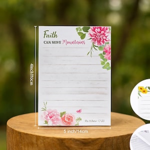 May include: A lined notepad with the text "Faith Can Move Mountains" and floral accents. The notepad is 10cm wide and 14cm long. Features pink and white flowers and the Bible verse Matthew 17:20.