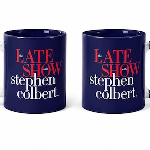 Könnte beinhalten: Zwei marineblaue Keramikbecher mit weißen Griffen. Jeder Becher trägt den Text "The Late Show with Stephen Colbert" in roter und weißer Schrift. Die Becher stehen vor einem weißen Hintergrund.