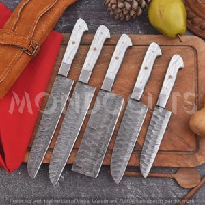 May include: A set of five kitchen knives with white handles and patterned Damascus steel blades. The knives are arranged in a row on a wooden cutting board. Each handle features a small decorative metal rivet. The blades display a distinct wavy pattern.