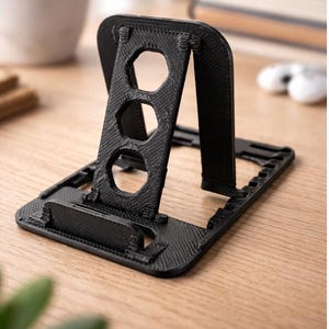 May include: A black, 3D-printed phone stand with a textured surface. The stand features a rectangular base, an adjustable back, and a support with three hexagonal cutouts. The stand is designed to hold a phone upright.