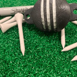 Peut inclure: Un support de balle de golf noir avec une balle de golf blanche et des rayures blanches. Le support a deux trous sur les côtés. Des tees de golf blancs et un marqueur Sharpie sont également visibles sur une surface verte.