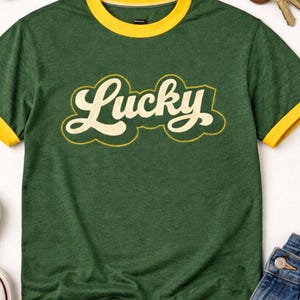 Pode incluir: Camiseta verde com gola e acabamento das mangas amarelas. A palavra "Lucky" é impressa no peito em uma fonte retrô creme com contorno amarelo. Óculos de sol, chaves e jeans são visíveis no fundo.